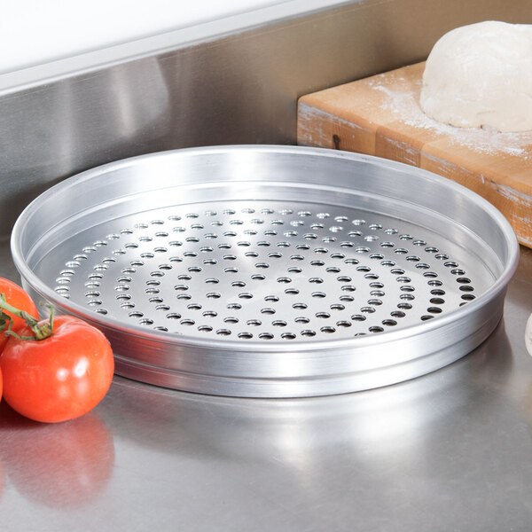 An American Metalcraft Super Perforated Pizza Pan on a metal surface with tomatoes and dough.