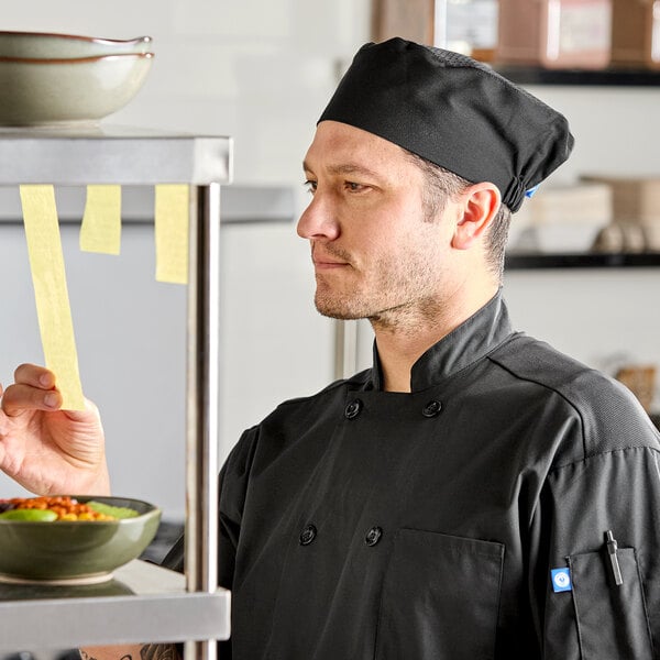 A black chef skull cap or pill box hat with a mesh top, worn by a chef in a kitchen setting.