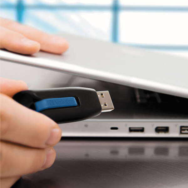 A hand holding a Verbatim Store 'n' Go V3 USB flash drive in front of a laptop.