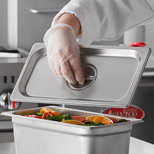 A person wearing gloves holding a Carlisle stainless steel lid over a food container.