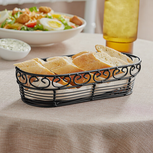 A Tablecraft black metal basket filled with bread on a table with a bowl of salad.