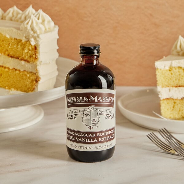 A bottle of Nielsen-Massey Madagascar Bourbon Vanilla Extract next to a slice of cake on a table.