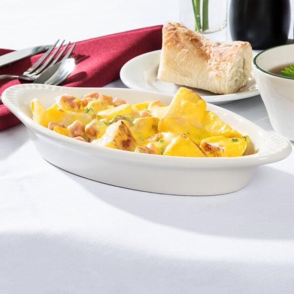 A white Tuxton oval dish with food in it on a table with a fork and knife.