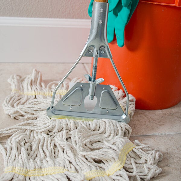 A Carlisle wooden stirrup style mop handle attached to a mop head in a mop bucket on the floor.