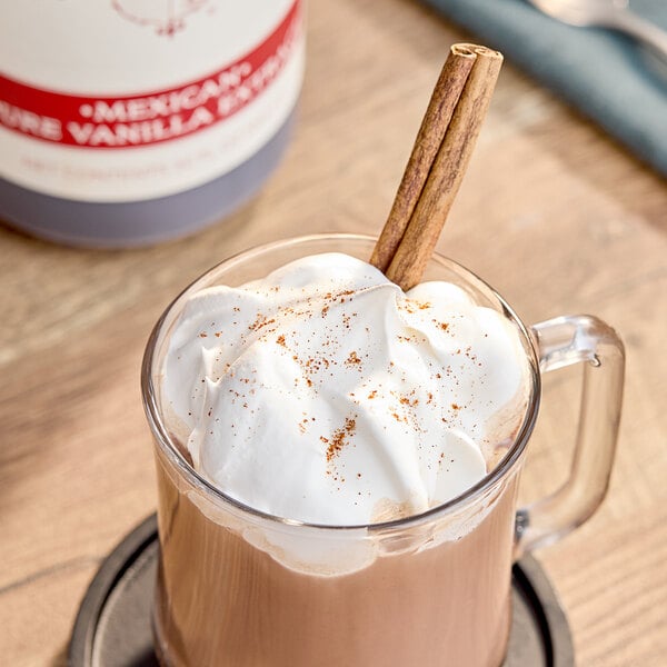 A glass mug of hot chocolate with cinnamon sticks and whipped cream made with Nielsen-Massey Mexican Vanilla Extract.