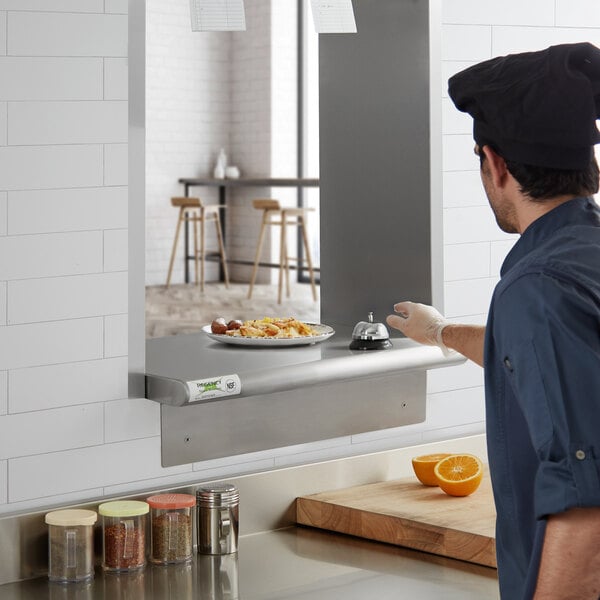 A man in a chef's uniform standing in front of a Regency stainless steel pass-through shelf with containers on it.