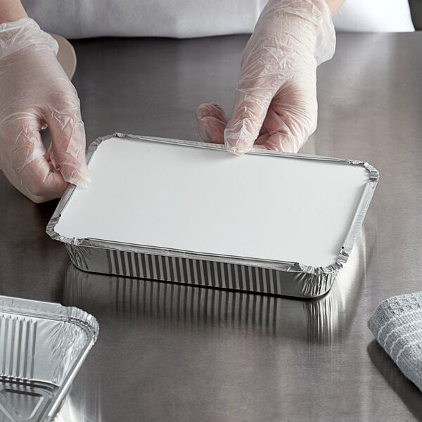 A person in gloves holding a white rectangular shallow oblong foil container with a board lid.