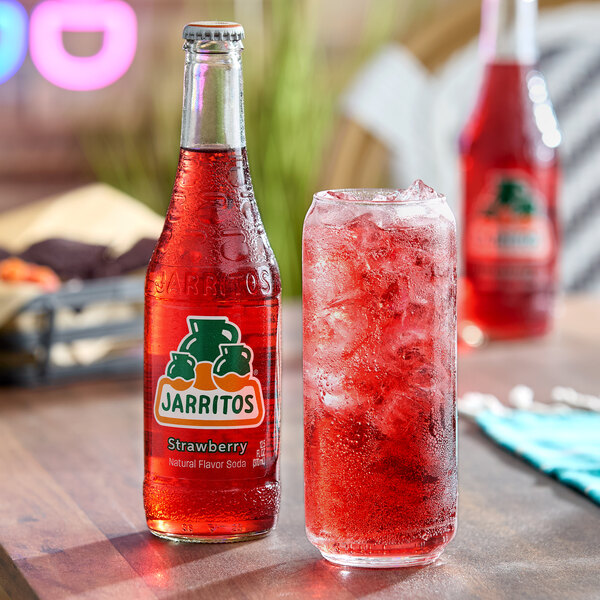 A bottle of Jarritos strawberry soda next to a glass filled with ice and red soda on a wooden table.
