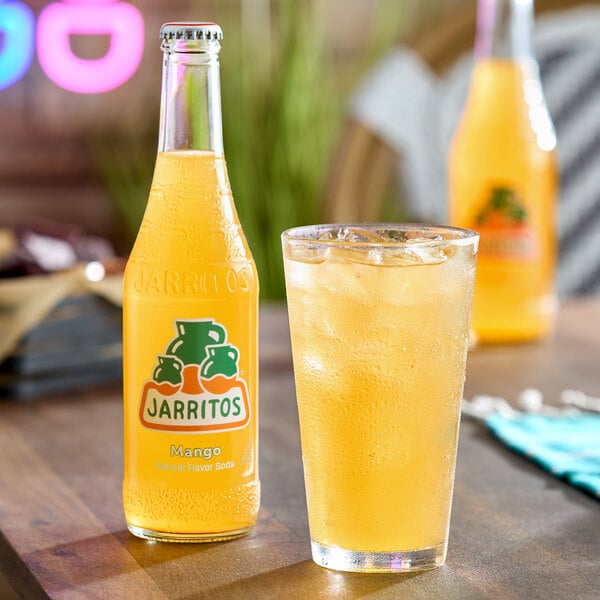 A bottle of Jarritos mango soda and a glass filled with ice and the same orange-colored beverage are placed on a wooden table.