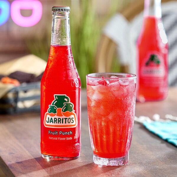 A bottle of Jarritos Fruit Punch soda is placed next to a glass filled with ice and the same red beverage on a table.