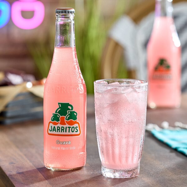 A bottle of pink Jarritos guava soda is placed next to a glass filled with ice and the same pink soda on a wooden table.