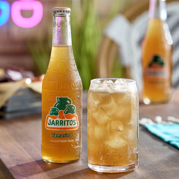 A bottle of Jarritos Tamarind soda next to a glass filled with ice and the same soda, both on a wooden table.