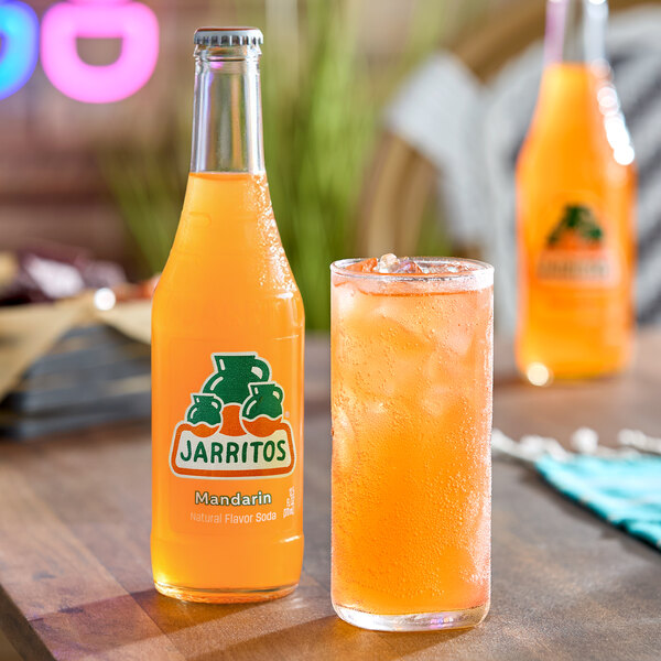 A bottle of orange Jarritos Mandarin soda is placed next to a glass filled with ice and the same orange beverage on a wooden table, with another bottle blurred in the background.