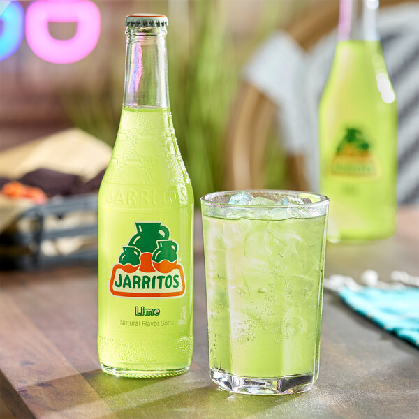 A glass bottle of Jarritos lime soda next to a glass filled with ice and lime soda on a wooden table.