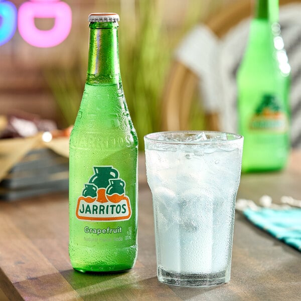 A green glass bottle of Jarritos Grapefruit soda is placed next to a glass filled with ice on a wooden table.