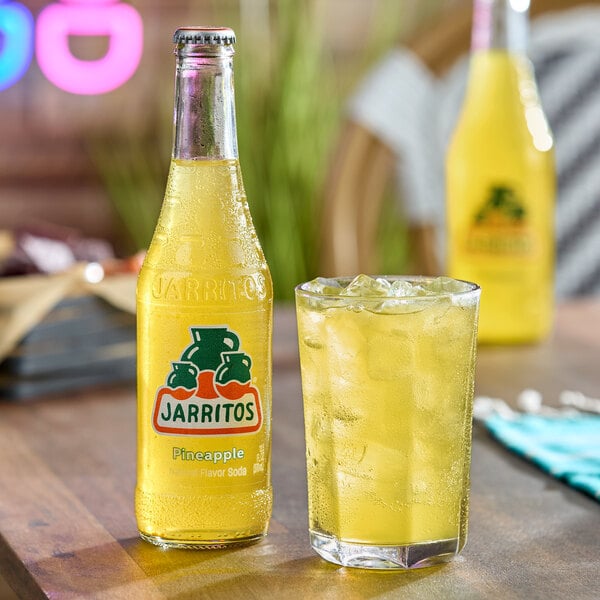 A bottle of yellow Jarritos soda and a glass filled with ice and the same yellow beverage are placed on a wooden table.