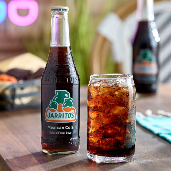 A glass bottle of Jarritos Mexican Cola and a glass filled with ice and cola are placed on a wooden table.