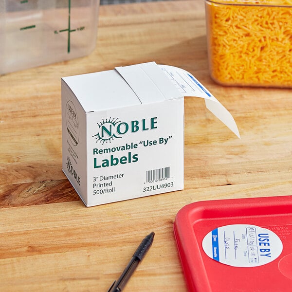 A white box of Noble Products 3" Use By Round Removable Labels on a table in a school kitchen.