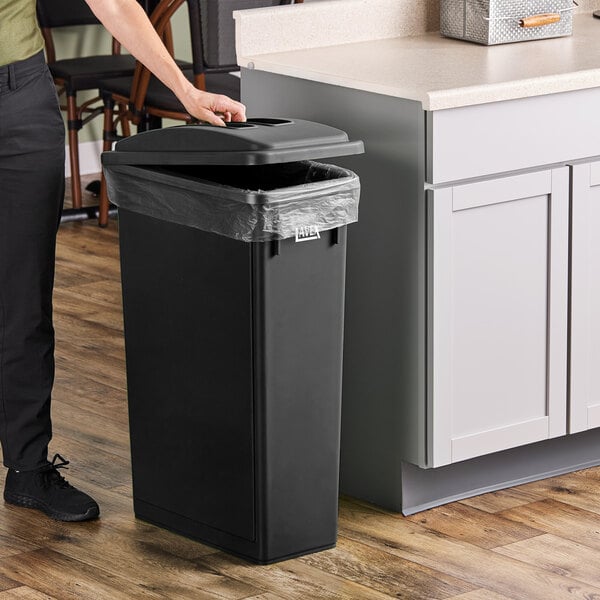 A woman standing next to a black Lavex slim rectangular trash can with a black flat lid.