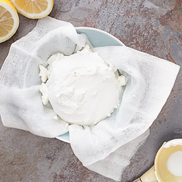 A bowl lined with white cheesecloth holding a mound of soft white cheese, with lemon slices and a measuring cup nearby.