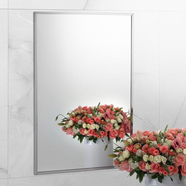 A Bobrick wall-mounted mirror reflecting a white vase with pink and white roses.
