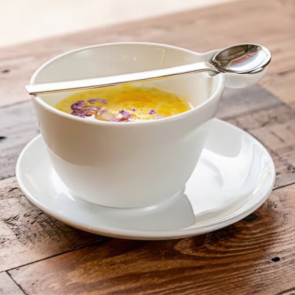 A white Front of the House Harmony porcelain cup with yellow liquid and a spoon on a wooden surface.