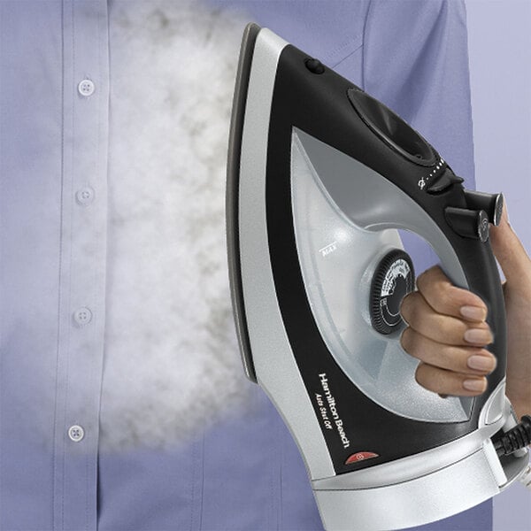 A person using a Hamilton Beach black and silver steam iron to iron a shirt.