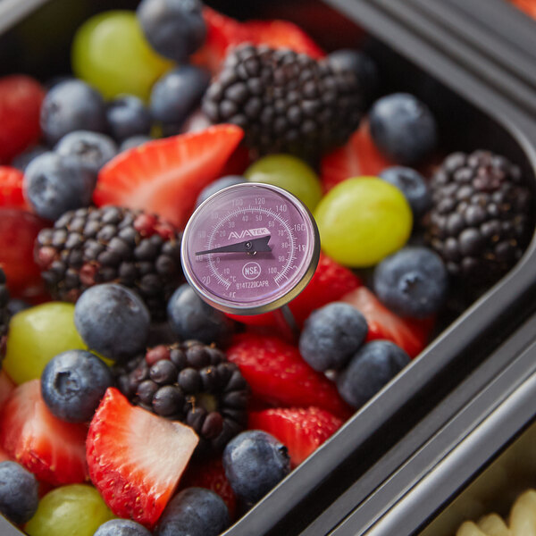 A purple dial probe thermometer inserted into a container of mixed fresh berries and grapes.