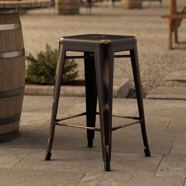 A Lancaster Table & Seating distressed copper metal outdoor backless counter height stool on a stone surface.