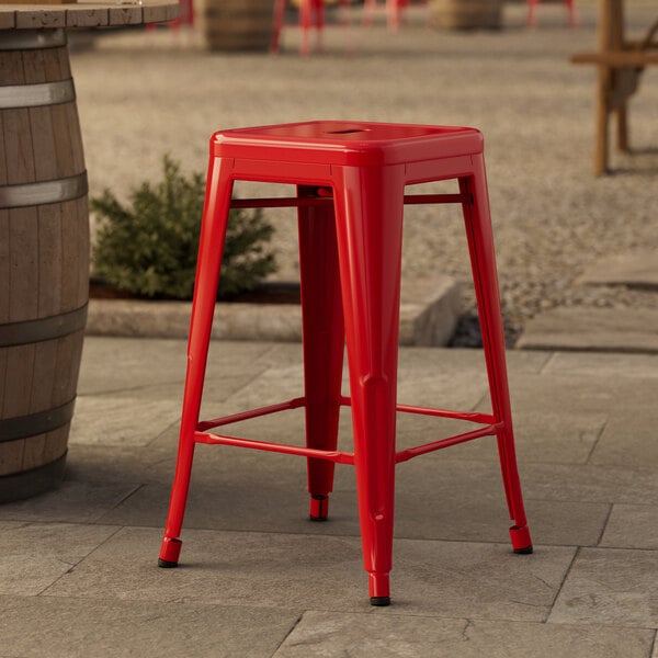 A red Lancaster Table & Seating outdoor backless counter height stool on a stone patio.