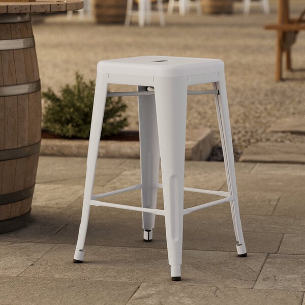 A white Lancaster Table & Seating outdoor backless counter height stool on a stone patio.