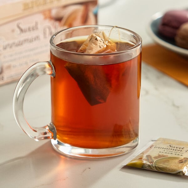A clear glass mug filled with brewed tea and a tea bag, with a packet of Bigelow Sweet Cinnamon Dolce Black Tea nearby.