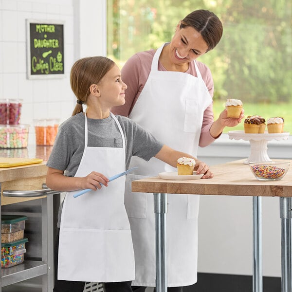 A woman and a girl in a school kitchen, both wearing white Uncommon Chef child bib aprons holding cupcakes.