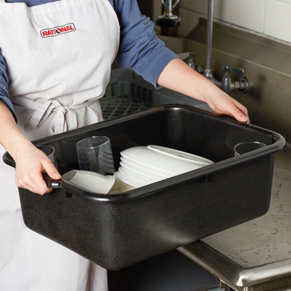 A woman in an apron holding a black Tablecraft bus tub on a counter.