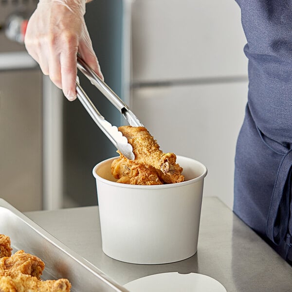 A person using tongs to put fried chicken in a white food bucket.