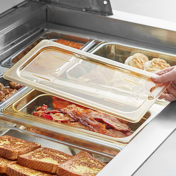 A person holding a Cambro 1/3 size amber high heat flat lid over a tray of food.