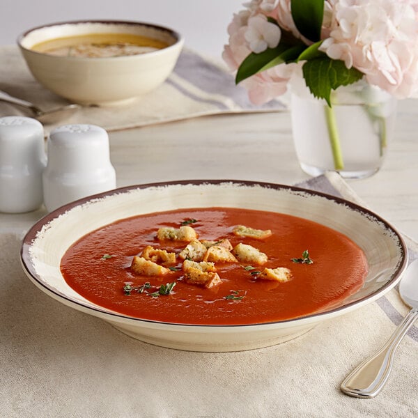A Carlisle white melamine soup bowl with a brown rim filled with soup and croutons with a spoon.