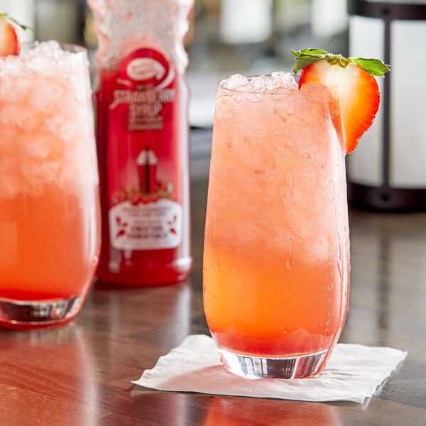A glass of pink strawberry drink with a strawberry on top on a napkin with a bottle of Master of Mixes Strawberry Syrup.