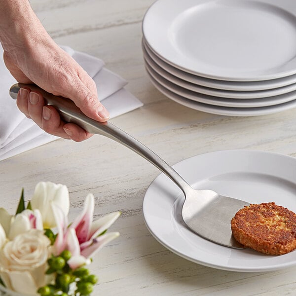A hand holding a Tablecraft Dalton stainless steel solid turner over a plate of food with a piece of food on it.