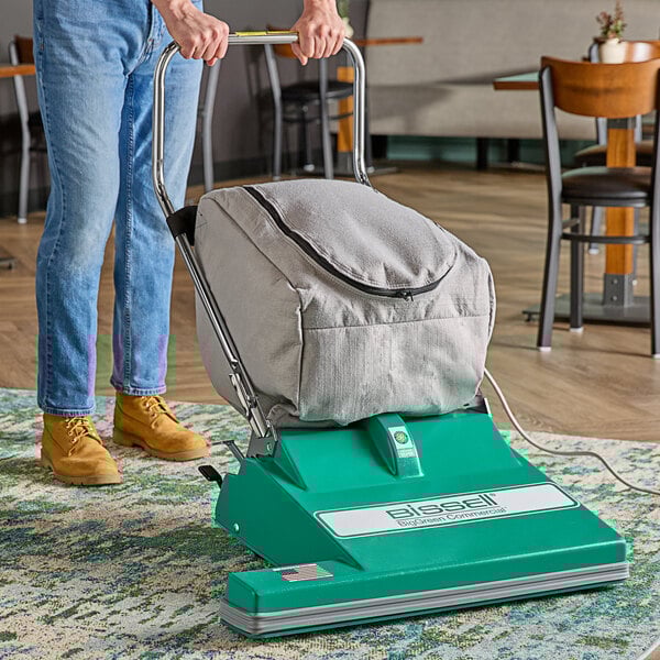 A person using a Bissell Commercial wide area vacuum cleaner with a green and metal handle to clean a rug.