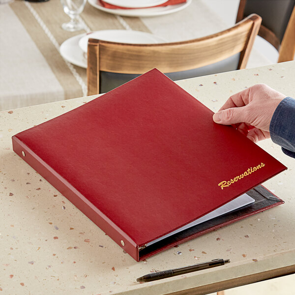 A hand holding a H. Risch wine faux leather reservation book with a glass of wine on a table.