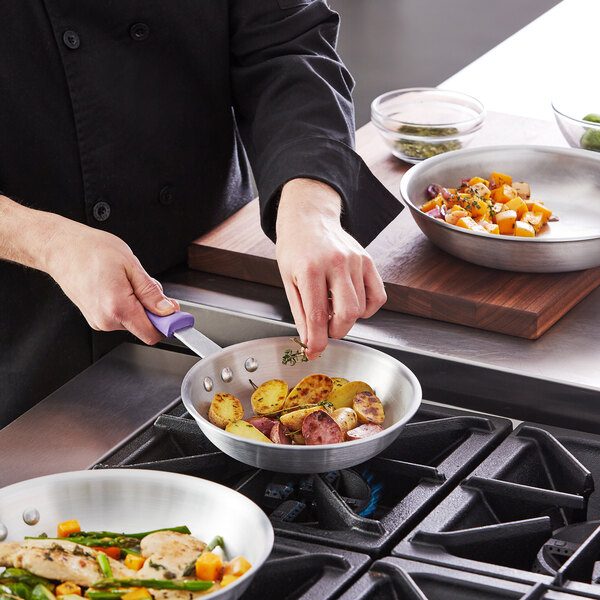 A person using a Choice aluminum fry pan with a purple silicone handle to cook food on a stove.