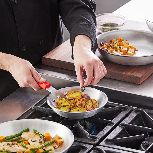 A person cooking food in a Choice aluminum fry pan with a red silicone handle.