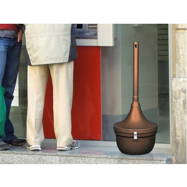 A man standing next to a copper Ex-Cell Kaiser Smokers' Oasis cigarette receptacle.