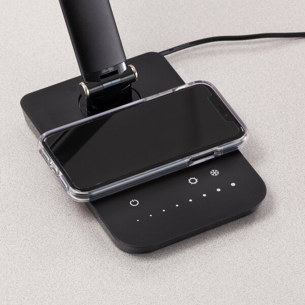 A black LED desk lamp with a multi-pivot adjustable arm, wireless charging pad, and USB port, shown charging a smartphone.
