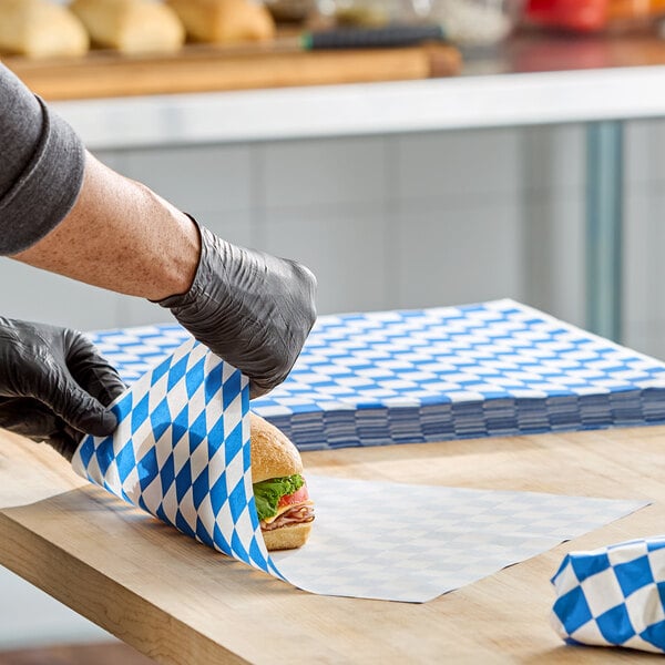 A stack of blue checkered deli sandwich wrap papers being used to wrap a sandwich on a wooden counter.