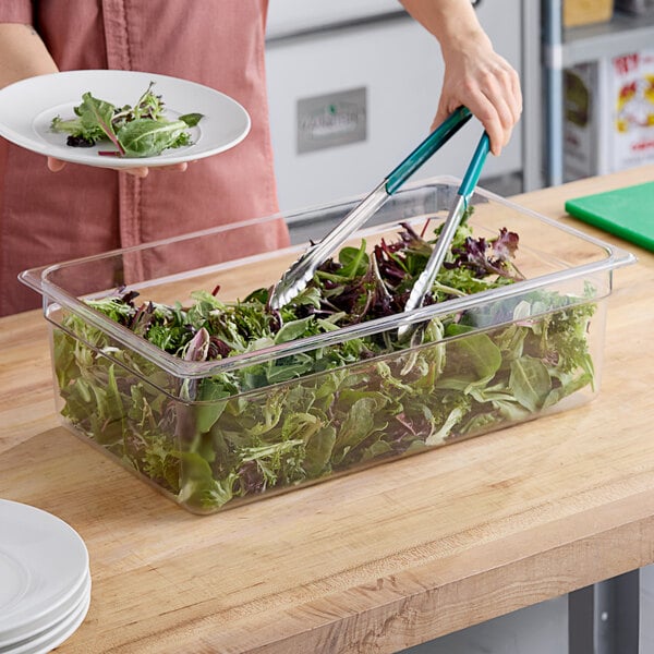 A woman using green tongs to serve salad from a Cambro clear plastic food pan on a salad bar counter.