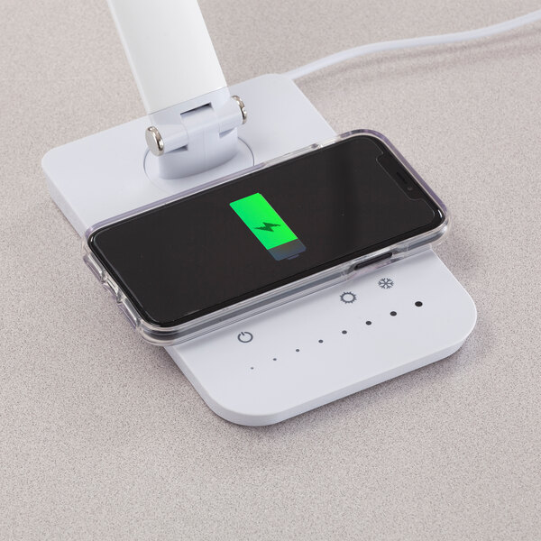 A white LED desk lamp with a multi-pivot adjustable arm, wireless charging pad, and USB port, shown charging a smartphone.