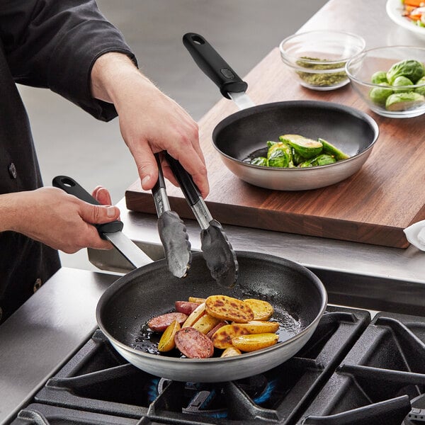 A person cooking meat and potatoes in Vollrath Wear-Ever frying pans on a stove.