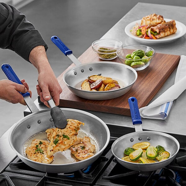 A man cooking brussels sprouts, potatoes, and chicken in Vollrath Wear-Ever frying pans with blue cool handles.